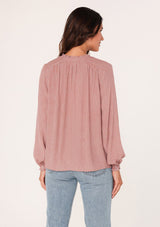 [Color: Mauve] A back facing image of a brunette model wearing a bohemian fall blouse in a mauve pink dot print. With long sleeves, a ruffled elastic wrist cuff, a self covered button front, a ruffled neckline, and a split v neckline.