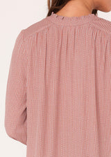 [Color: Mauve] A close up back facing image of a brunette model wearing a bohemian fall blouse in a mauve pink dot print. With long sleeves, a ruffled elastic wrist cuff, a self covered button front, a ruffled neckline, and a split v neckline.