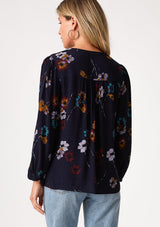 [Color: Navy/Mustard] A back facing image of a blonde model wearing a bohemian fall blouse in a blue floral print. With long sleeves, a v neckline, a self covered button front, and a relaxed fit. 