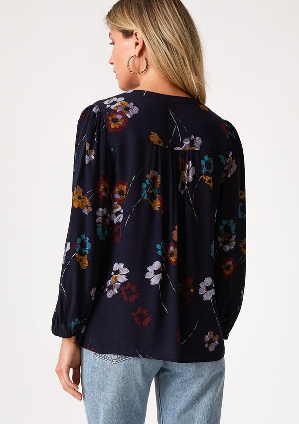 [Color: Navy/Mustard] A back facing image of a blonde model wearing a bohemian fall blouse in a blue floral print. With long sleeves, a v neckline, a self covered button front, and a relaxed fit. 