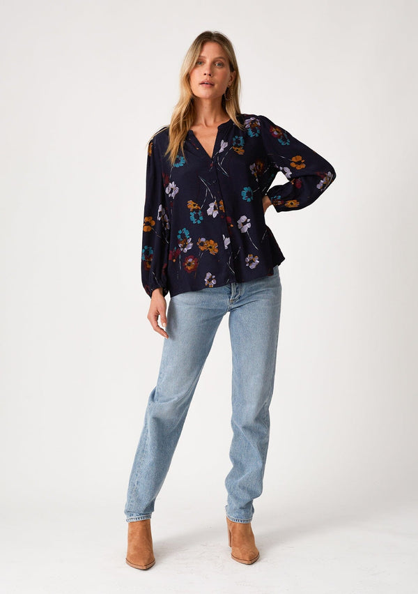 [Color: Navy/Mustard] A front facing image of a blonde model wearing a bohemian fall blouse in a blue floral print. With long sleeves, a v neckline, a self covered button front, and a relaxed fit. 