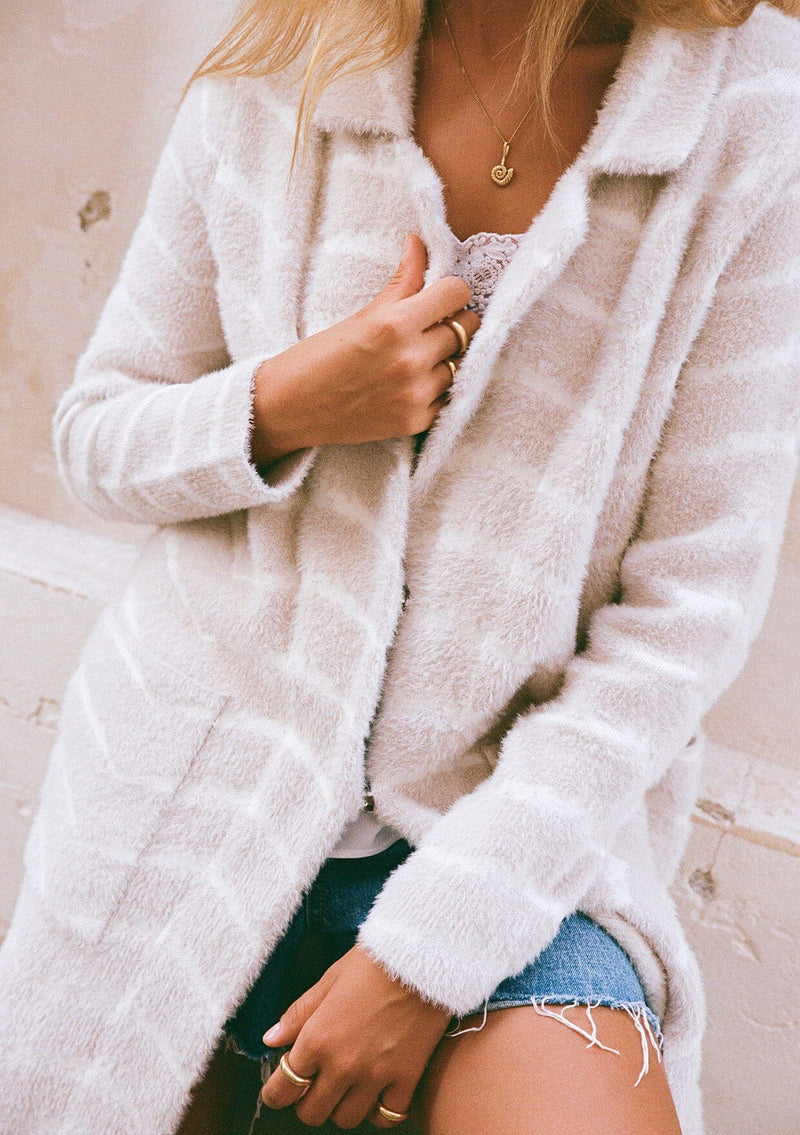 [Color: Sand/Cream] A close up front facing image of a blonde model wearing a soft and fuzzy sweater coat in an ivory and white chevron design. With a snap button front, side pockets, and a classic notched lapel.