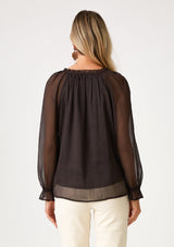 [Color: Chocolate] A back facing image of a blonde model wearing a bohemian fall blouse in a dark brown sheer chiffon. With long raglan sleeves, ruffled elastic wrist cuffs, a ruffled v neckline, and a self covered button front. 