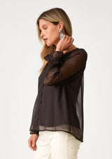 [Color: Chocolate] A side facing image of a blonde model wearing a bohemian fall blouse in a dark brown sheer chiffon. With long raglan sleeves, ruffled elastic wrist cuffs, a ruffled v neckline, and a self covered button front. 