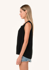 [Color: Black] A side facing image of a blonde model wearing a lightweight classic sleeveless blouse in black. With a ruffled neckline, a split v neckline with ties, and a relaxed fit.