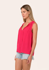 [Color: Watermelon] A side facing image of a blonde model wearing a lightweight classic sleeveless blouse in bright pink. With a ruffled neckline, a split v neckline with ties, and a relaxed fit.