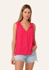 [Color: Watermelon] A front facing image of a blonde model wearing a lightweight classic sleeveless blouse in bright pink. With a ruffled neckline, a split v neckline with ties, and a relaxed fit.
