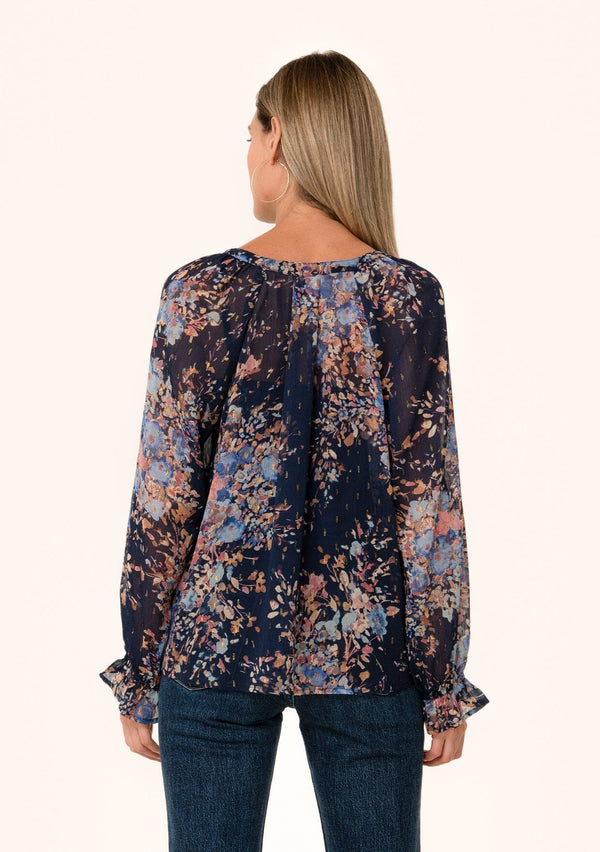 [Color: Navy/Dusty Blue] A back facing image of a blonde model wearing a bohemian fall blouse in a blue floral print. Designed in a metallic clip dot chiffon, with sheer long sleeves, ruffled elastic wrist cuffs, a button loop trimmed v neckline, and a self covered button front. 