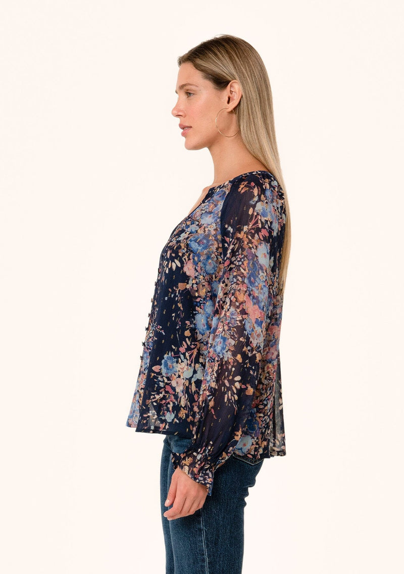 [Color: Navy/Dusty Blue] A side facing image of a blonde model wearing a bohemian fall blouse in a blue floral print. Designed in a metallic clip dot chiffon, with sheer long sleeves, ruffled elastic wrist cuffs, a button loop trimmed v neckline, and a self covered button front. 