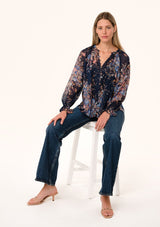 [Color: Navy/Dusty Blue] A front facing image of a blonde model sitting on a stool wearing a bohemian fall blouse in a blue floral print. Designed in a metallic clip dot chiffon, with sheer long sleeves, ruffled elastic wrist cuffs, a button loop trimmed v neckline, and a self covered button front. 