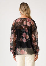 [Color: Black/Rose] A back facing image of a blonde model wearing a bohemian holiday blouse in a black and pink sheer floral chiffon. With voluminous long sleeves, a v neckline, and a self covered button front. 