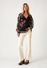 [Color: Black/Rose] A full body front facing image of a blonde model wearing a bohemian holiday blouse in a black and pink sheer floral chiffon. With voluminous long sleeves, a v neckline, and a self covered button front. 