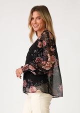 [Color: Black/Rose] A side facing image of a blonde model wearing a bohemian holiday blouse in a black and pink sheer floral chiffon. With voluminous long sleeves, a v neckline, and a self covered button front. 