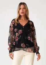 [Color: Black/Rose] A front facing image of a blonde model wearing a bohemian holiday blouse in a black and pink sheer floral chiffon. With voluminous long sleeves, a v neckline, and a self covered button front. 
