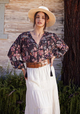 [Color: Brown/Dusty Lilac] A half body front facing image of a brunette model standing outside wearing a bohemian fall blouse in a brown and dusty purple floral print. With three quarter length sleeves, a self covered button front, a split v neckline with tassel ties, and a relaxed fit.