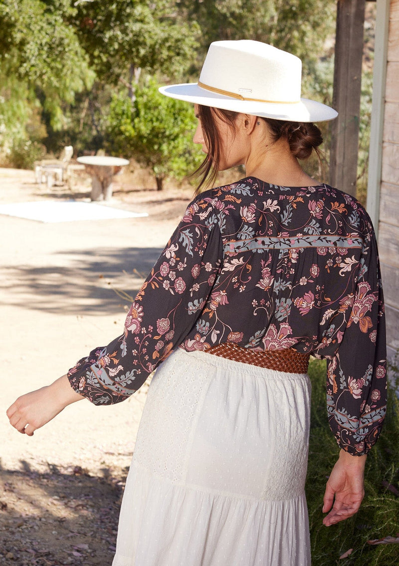 [Color: Brown/Dusty Lilac] A half body back facing image of a brunette model standing outside wearing a bohemian fall blouse in a brown and dusty purple floral print. With three quarter length sleeves, a self covered button front, a split v neckline with tassel ties, and a relaxed fit.