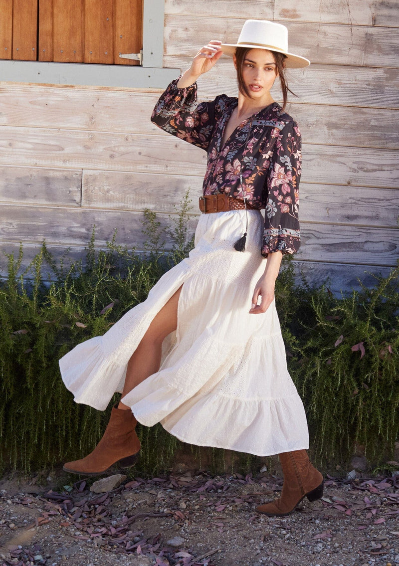 [Color: Natural] A side facing image of a brunette model wearing a bohemian off white cotton maxi skirt with embroidered detail throughout.