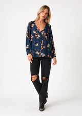 [Color: Teal/Dusty Blush] A full body front facing image of a blonde model wearing a bohemian fall blouse in a teal blue floral print. With voluminous long raglan sleeves, a v neckline, a smocked shoulder detail, and a self covered button front. 