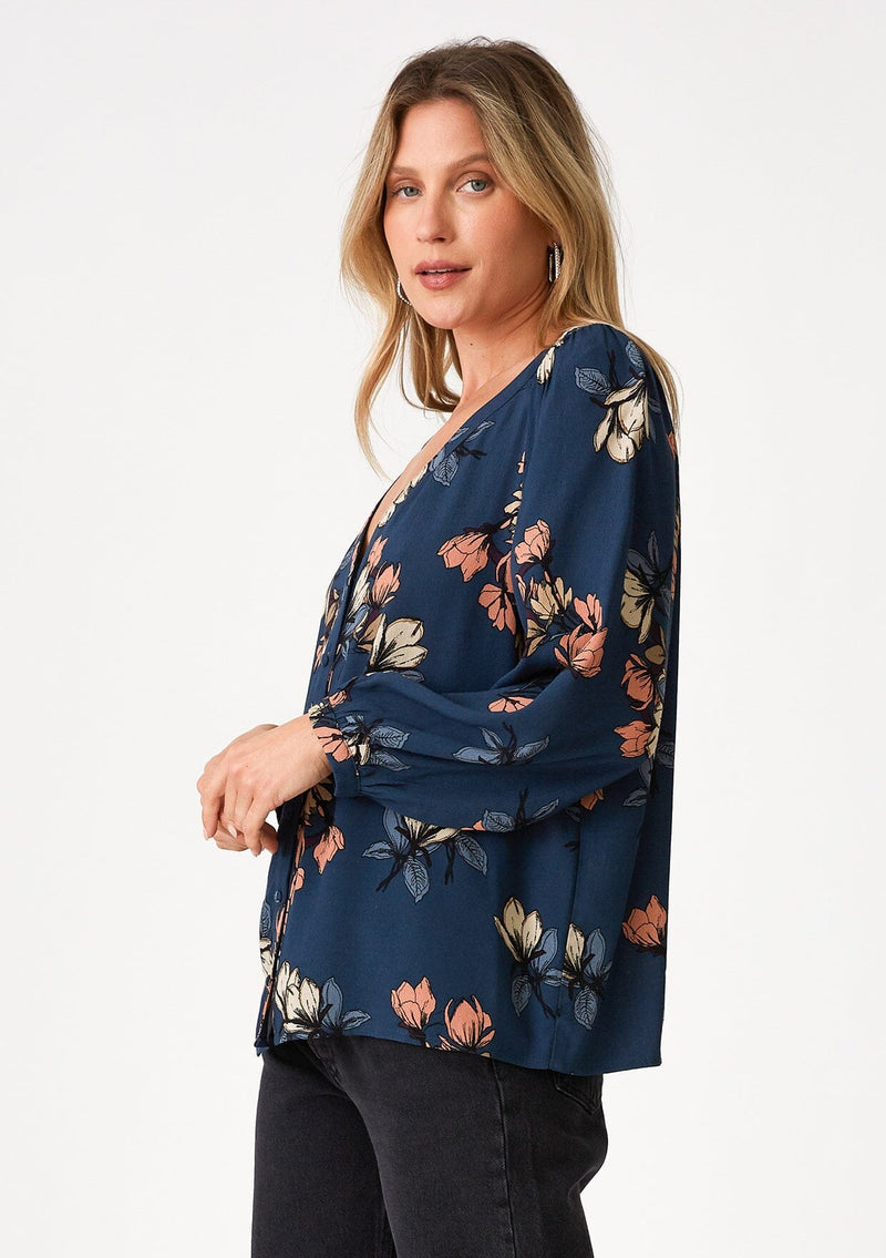 [Color: Teal/Dusty Blush] A side facing image of a blonde model wearing a bohemian fall blouse in a teal blue floral print. With voluminous long raglan sleeves, a v neckline, a smocked shoulder detail, and a self covered button front. 