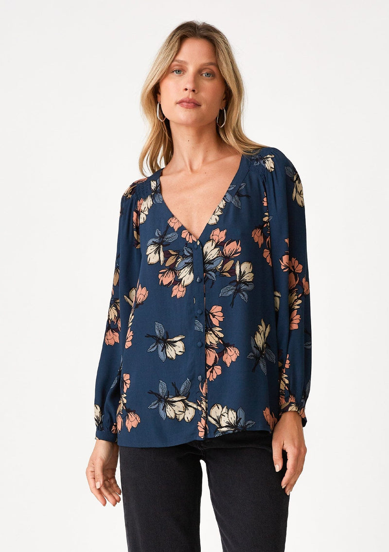 [Color: Teal/Dusty Blush] A front facing image of a blonde model wearing a bohemian fall blouse in a teal blue floral print. With voluminous long raglan sleeves, a v neckline, a smocked shoulder detail, and a self covered button front. 