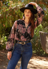 [Color: Black/Dusty Rose] A half body front facing image of a brunette model standing outside wearing a bohemian chiffon blouse designed in a black and pink floral print. With metallic details throughout, a surplice v neckline, long sleeves, and an elastic waist at the front.