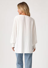[Color: Cream] A back facing image of a blonde model wearing a bohemian off white blouse. With long sleeves, side vents, smocked ruffled wrist cuffs, a button loop trimmed v neckline, and a self covered button front. 