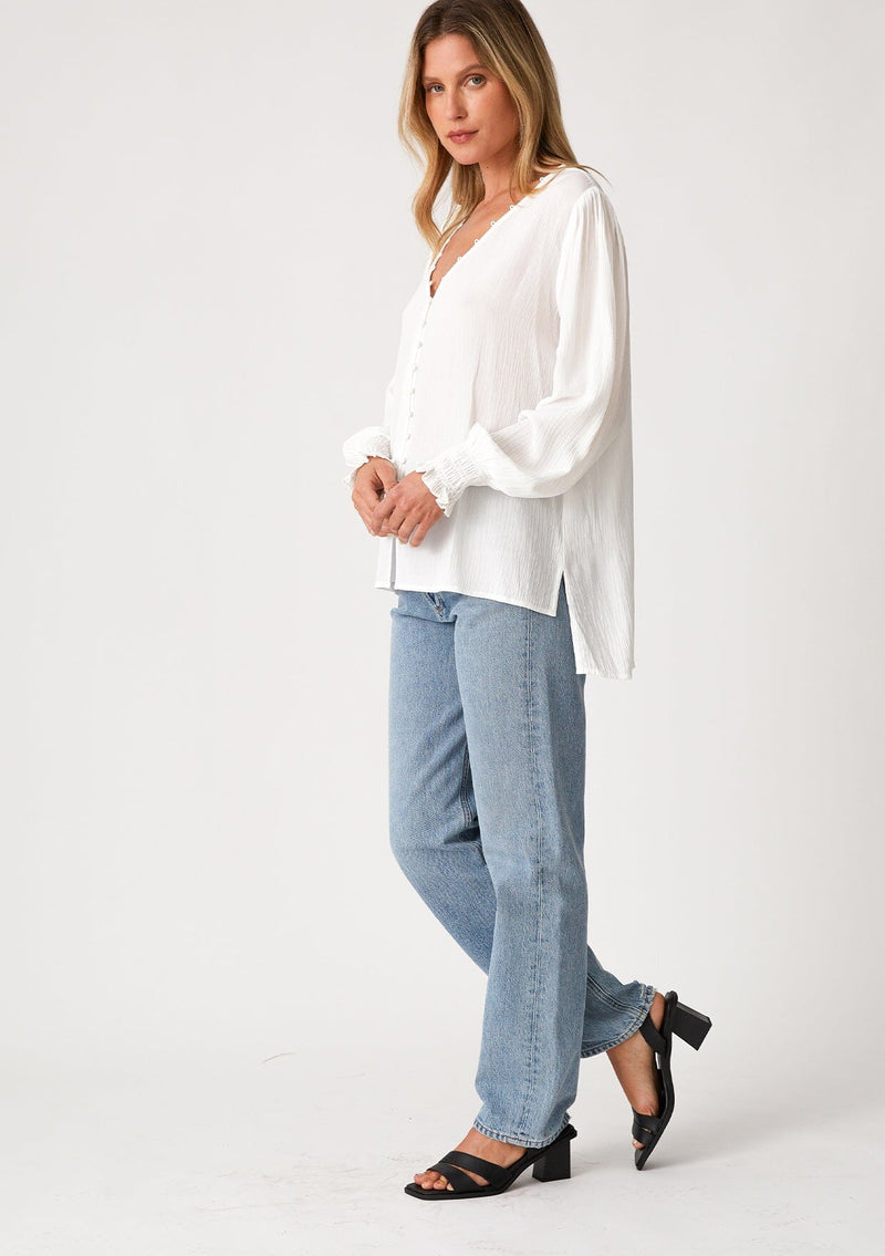 [Color: Cream] A side facing image of a blonde model wearing a bohemian off white blouse. With long sleeves, side vents, smocked ruffled wrist cuffs, a button loop trimmed v neckline, and a self covered button front. 