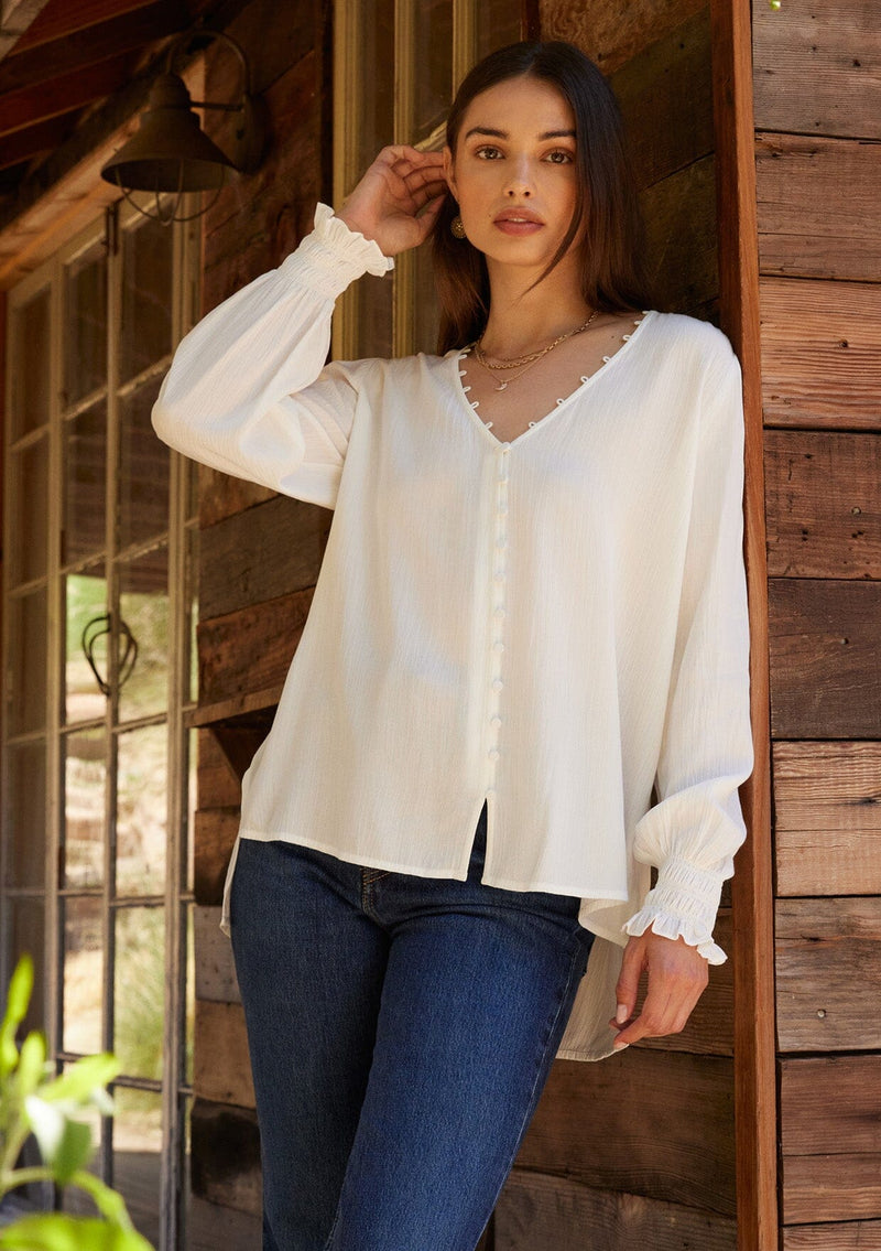 [Color: Cream] A half body front facing image of a brunette model wearing a bohemian off white blouse. With long sleeves, side vents, smocked ruffled wrist cuffs, a button loop trimmed v neckline, and a self covered button front.