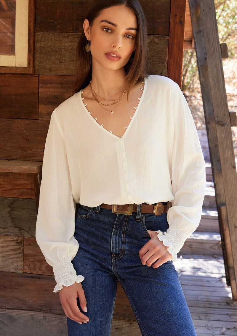 [Color: Cream] A front facing image of a brunette model wearing a bohemian off white blouse. With long sleeves, side vents, smocked ruffled wrist cuffs, a button loop trimmed v neckline, and a self covered button front.