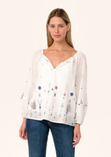 [Color: Natural/Olive] A front facing image of a blonde model wearing a white bohemian cotton summer blouse with colorful embroidered details. With long sleeves, a split v neckline with tassel ties, and a relaxed, flowy fit.