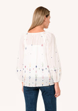 [Color: Natural/Olive] A back facing image of a blonde model wearing a white bohemian cotton summer blouse with colorful embroidered details. With long sleeves, a split v neckline with tassel ties, and a relaxed, flowy fit. 