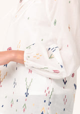 [Color: Natural/Olive] A close up side facing image of a blonde model wearing a white bohemian cotton summer blouse with colorful embroidered details. With long sleeves, a split v neckline with tassel ties, and a relaxed, flowy fit. 
