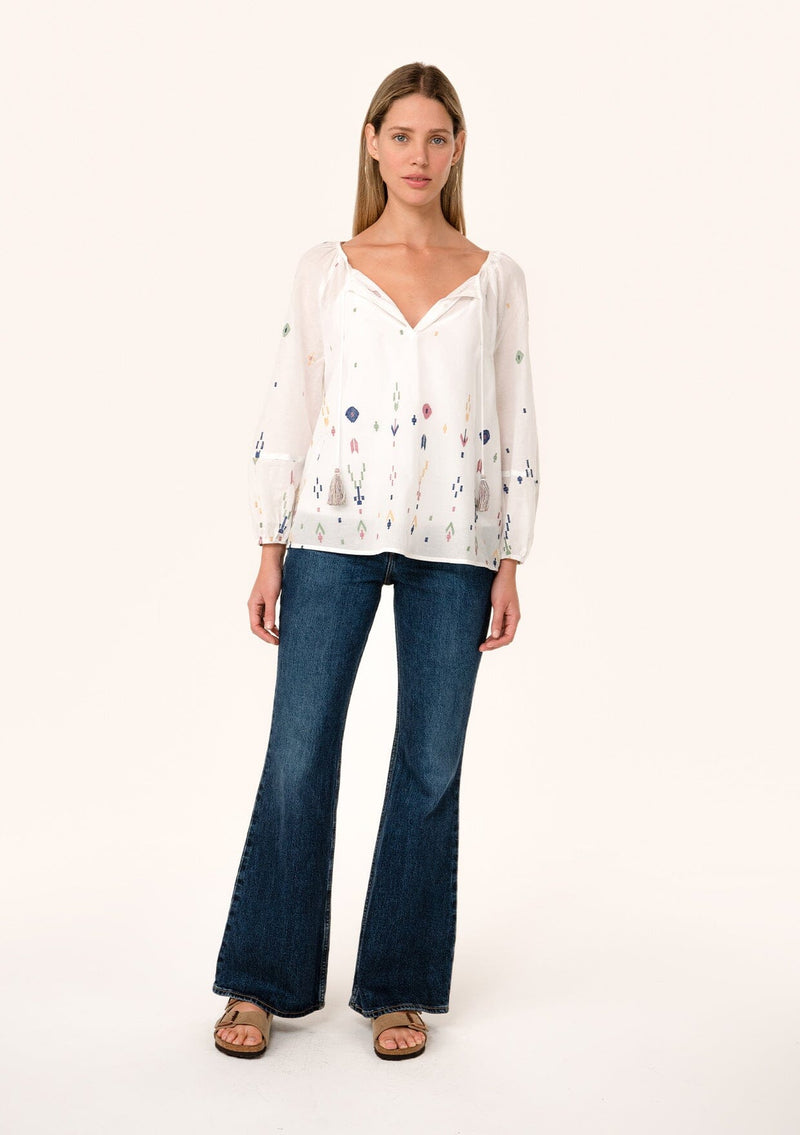 [Color: Natural/Olive] A full body front facing image of a blonde model wearing a white bohemian cotton summer blouse with colorful embroidered details. With long sleeves, a split v neckline with tassel ties, and a relaxed, flowy fit. 