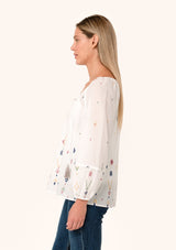 [Color: Natural/Olive] A side facing image of a blonde model wearing a white bohemian cotton summer blouse with colorful embroidered details. With long sleeves, a split v neckline with tassel ties, and a relaxed, flowy fit. 