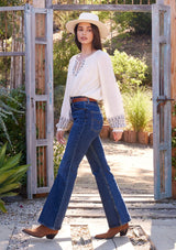 [Color: Natural] A side facing image of a brunette model standing outside wearing a bohemian blouse with embroidered details. With voluminous long sleeves, a split v neckline with tassel ties, a smocked neckline, and a relaxed fit.