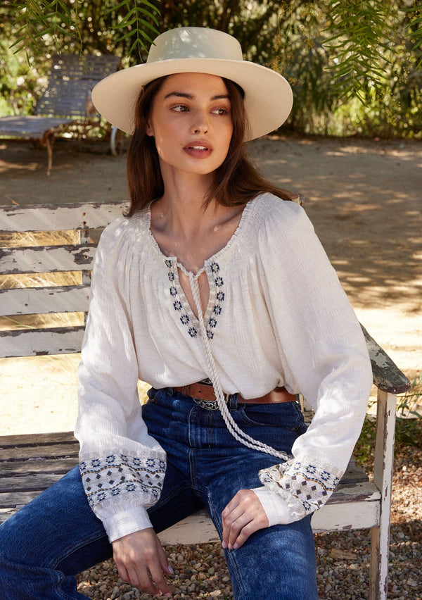[Color: Natural] A front facing image of a brunette model sitting outside wearing a bohemian blouse with embroidered details. With voluminous long sleeves, a split v neckline with tassel ties, a smocked neckline, and a relaxed fit.