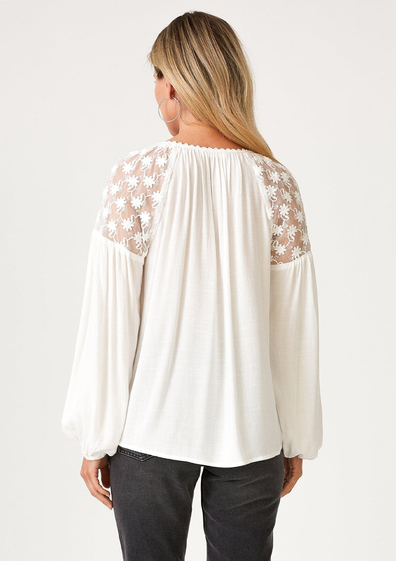 [Color: Ivory] A back facing image of a blonde model wearing a white bohemian holiday blouse with long sleeves, a split v neckline with ties, and sheer embroidered mesh detail along the shoulders. 