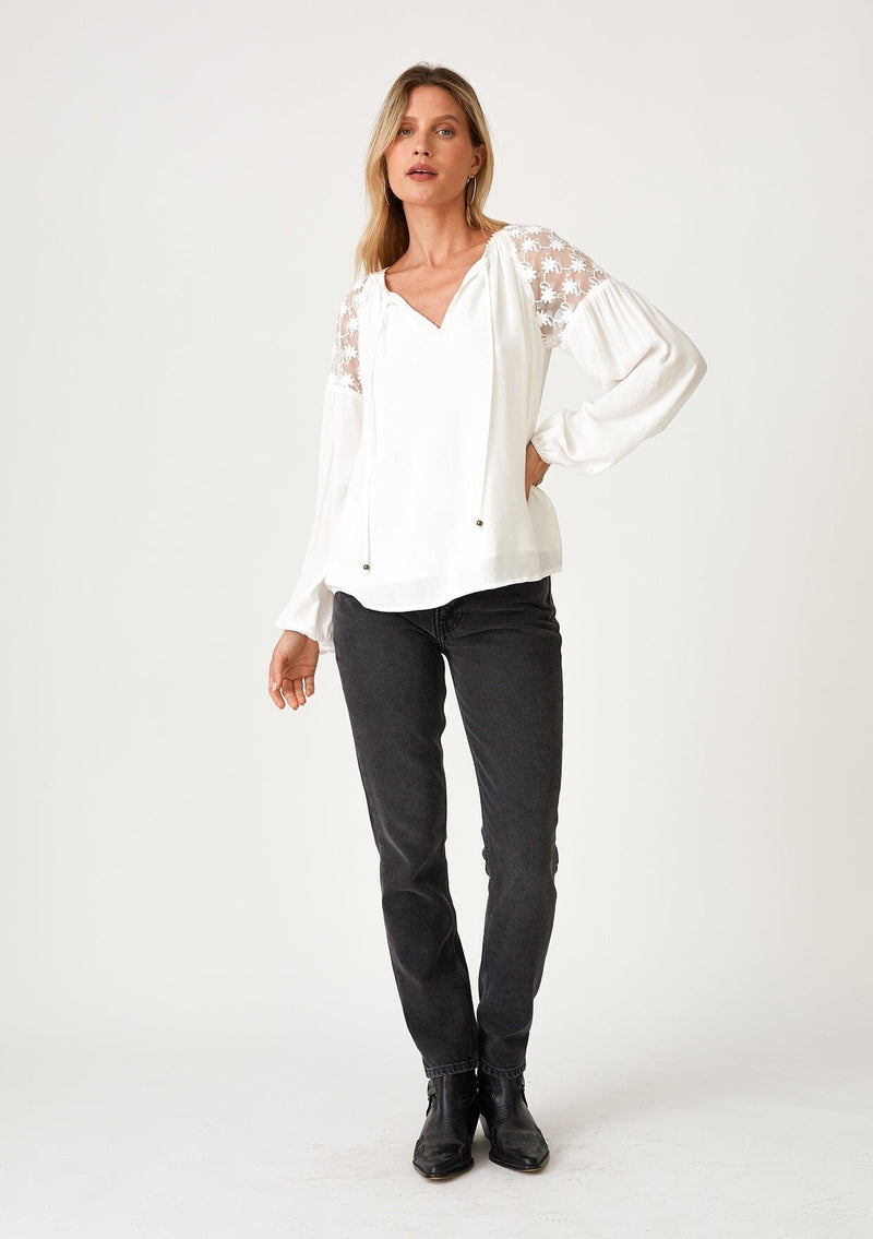 [Color: Ivory] A full body front facing image of a blonde model wearing a white bohemian holiday blouse with long sleeves, a split v neckline with ties, and sheer embroidered mesh detail along the shoulders. 