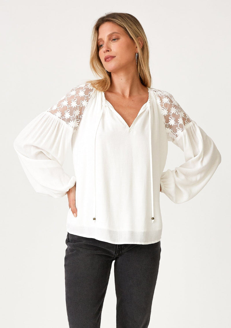 [Color: Ivory] A front facing image of a blonde model wearing a white bohemian holiday blouse with long sleeves, a split v neckline with ties, and sheer embroidered mesh detail along the shoulders. 
