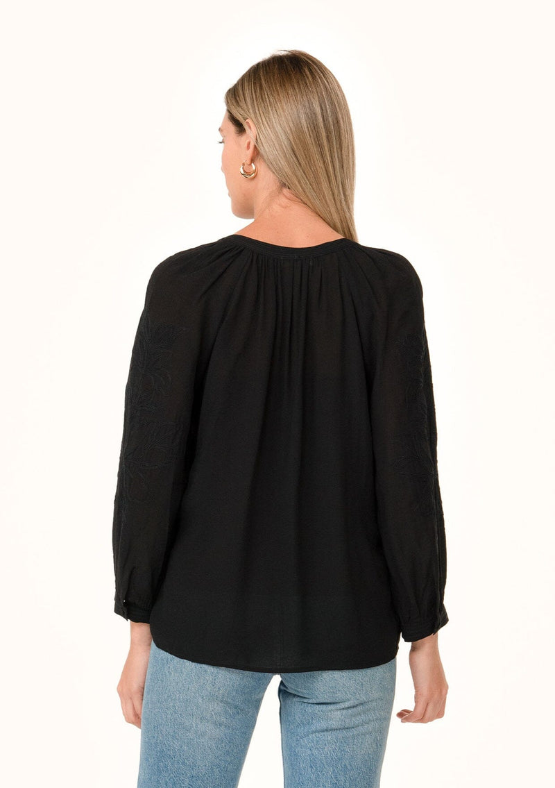 [Color: Black] A back facing image of a blonde model wearing a black bohemian fall blouse. With a v neckline, long sleeves, and embroidered detail.