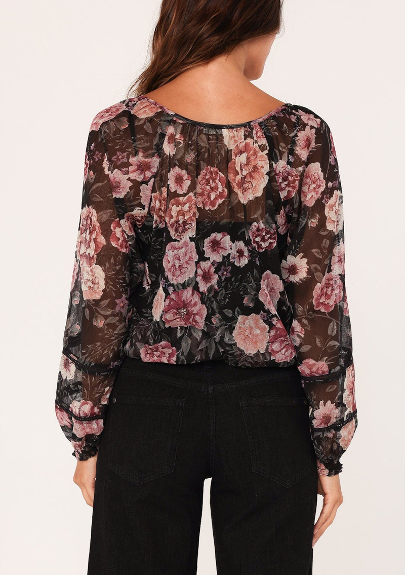 [Color: Black/Rose] A back facing image of a brunette model wearing a sheer chiffon bohemian blouse in a black and pink floral print. With sheer long sleeves, a split v neckline with ties, a smocked elastic waist, and lattice trim. 