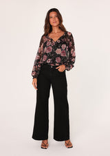 [Color: Black/Rose] A full body front facing image of a brunette model wearing a sheer chiffon bohemian blouse in a black and pink floral print. With sheer long sleeves, a split v neckline with ties, a smocked elastic waist, and lattice trim. 