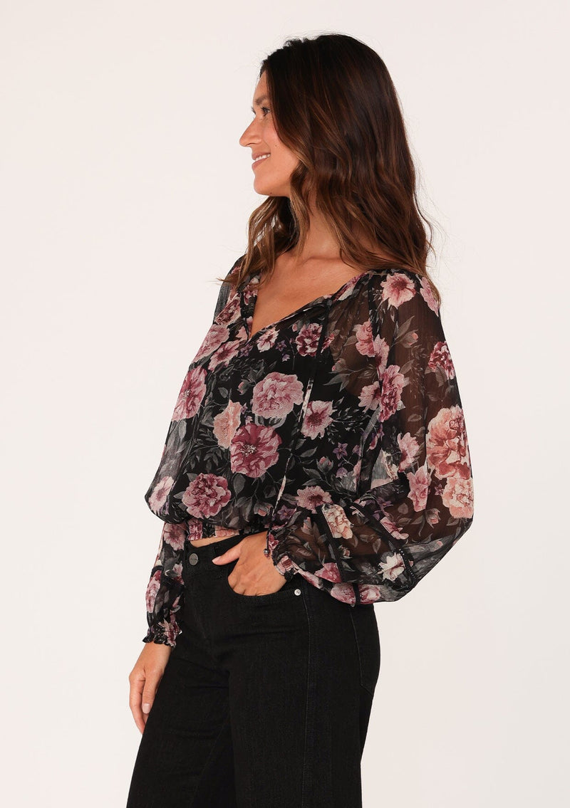 [Color: Black/Rose] A side facing image of a brunette model wearing a sheer chiffon bohemian blouse in a black and pink floral print. With sheer long sleeves, a split v neckline with ties, a smocked elastic waist, and lattice trim. 