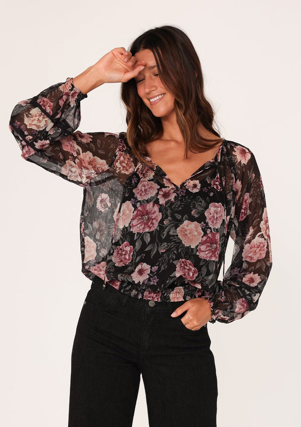 [Color: Black/Rose] A front facing image of a brunette model wearing a sheer chiffon bohemian blouse in a black and pink floral print. With sheer long sleeves, a split v neckline with ties, a smocked elastic waist, and lattice trim. 