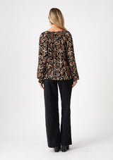 [Color: Black/Taupe] A back facing image of a blonde model wearing a flowy bohemian blouse in a brown bohemian print. With voluminous long raglan sleeves, a split v neckline with ties, and a self covered button front. 