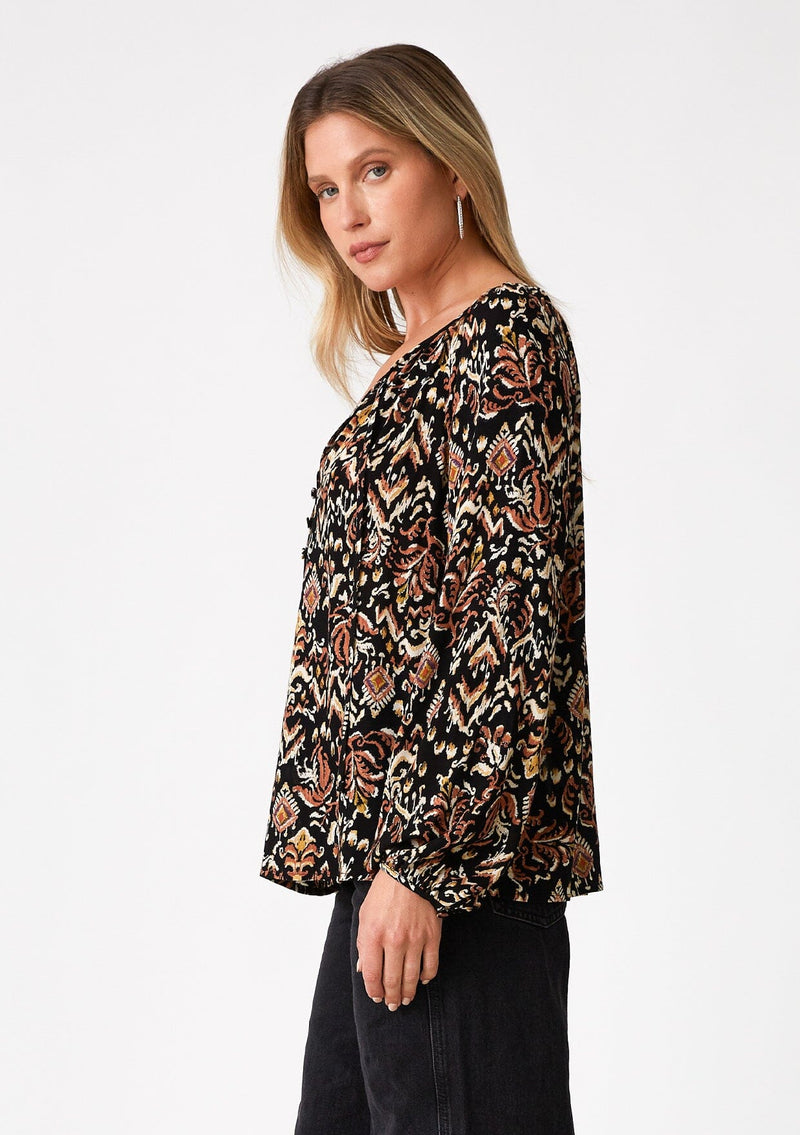 [Color: Black/Taupe] A side facing image of a blonde model wearing a flowy bohemian blouse in a brown bohemian print. With voluminous long raglan sleeves, a split v neckline with ties, and a self covered button front. 