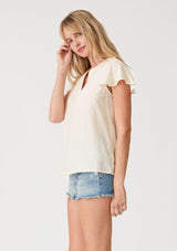 [Color: Cream] A side facing image of a blonde model wearing a lightweight bohemian resort top  in cream. With short flutter sleeves, a split v neckline with embroidered top stitch details, and a relaxed fit. 
