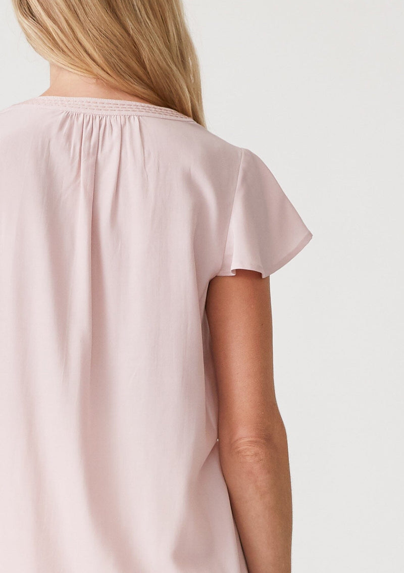 [Color: Dusty Pink] A close up back facing image of a blonde model wearing a lightweight bohemian resort top  in dusty pink. With short flutter sleeves, a split v neckline with embroidered top stitch details, and a relaxed fit. 