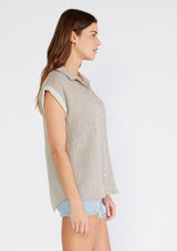 [Color: Taupe] A side facing image of a brunette model wearing a light brown short sleeve shirt crafted from soft cotton gauze. With a button up front, a collared neckline, a front patch pocket, and short contrast cap sleeves. 