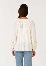 [Color: Cream/Natural] A back facing image of a brunette model wearing a cream bohemian fall blouse with embroidered details and sparkly metallic thread. With long raglan sleeves, a split v neckline with tassel ties, lattice trim, and a relaxed fit.